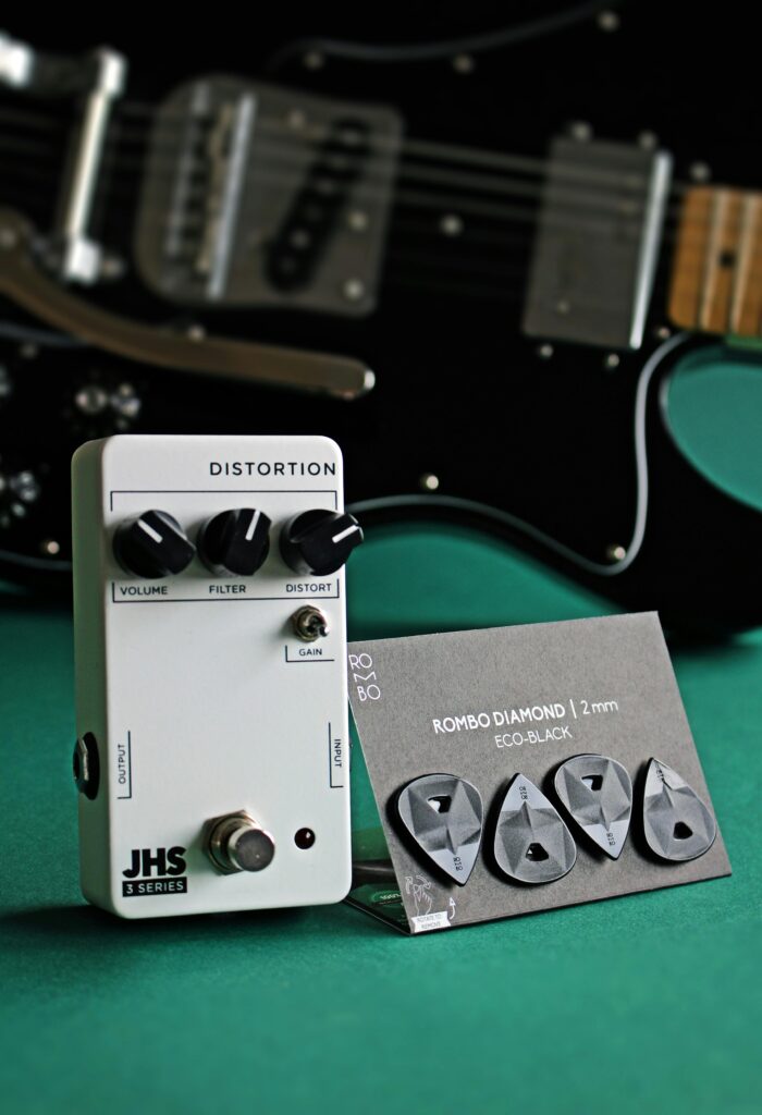 Distortion pedal and guitar picks beside electric guitar on green surface.